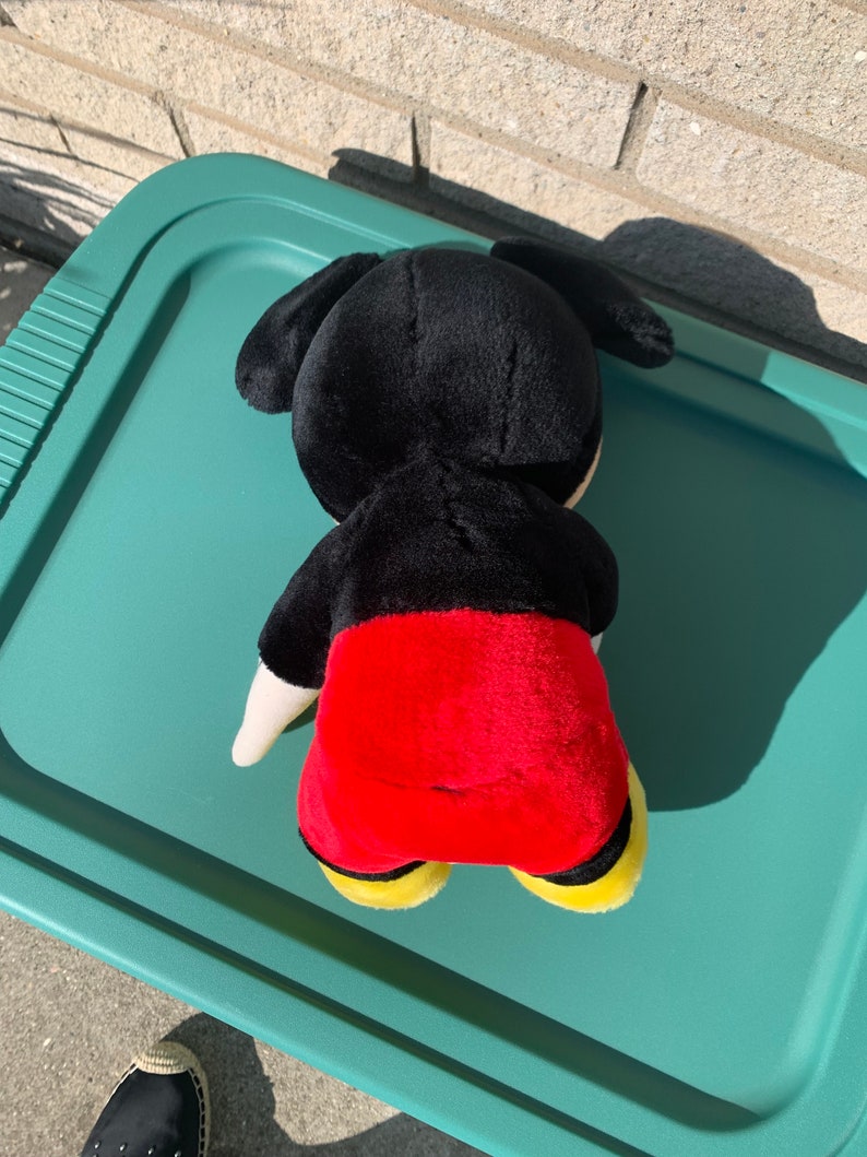 Puede incluir: Un juguete de peluche negro y rojo de Mickey Mouse. El juguete est&aacute; acostado boca abajo con la cabeza girada hacia un lado. El juguete lleva zapatos amarillos.