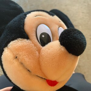 Puede incluir: Primer plano de un juguete de peluche de Mickey Mouse con orejas negras, nariz negra y boca roja. El juguete tiene una cara marr&oacute;n claro con un ojo blanco y una pupila negra.