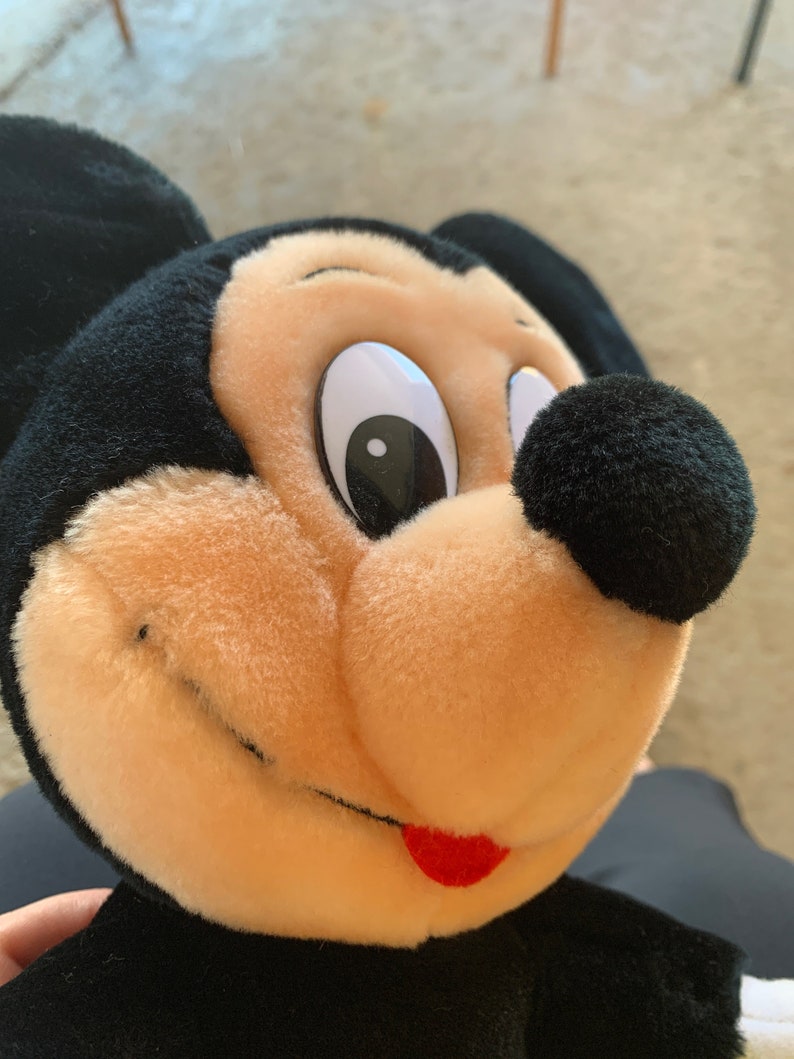 Puede incluir: Primer plano de un juguete de peluche de Mickey Mouse con orejas negras, nariz negra y boca roja. El juguete tiene una cara marr&oacute;n claro con un ojo blanco y una pupila negra.