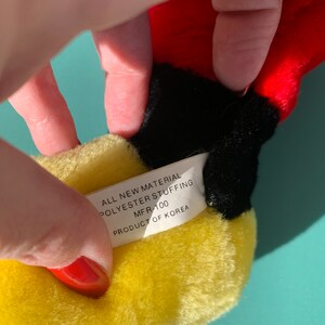 Puede incluir: Primer plano de un juguete de peluche amarillo con una etiqueta blanca que dice "All New Material Polyester Stuffing MFR-100 Product of Korea".