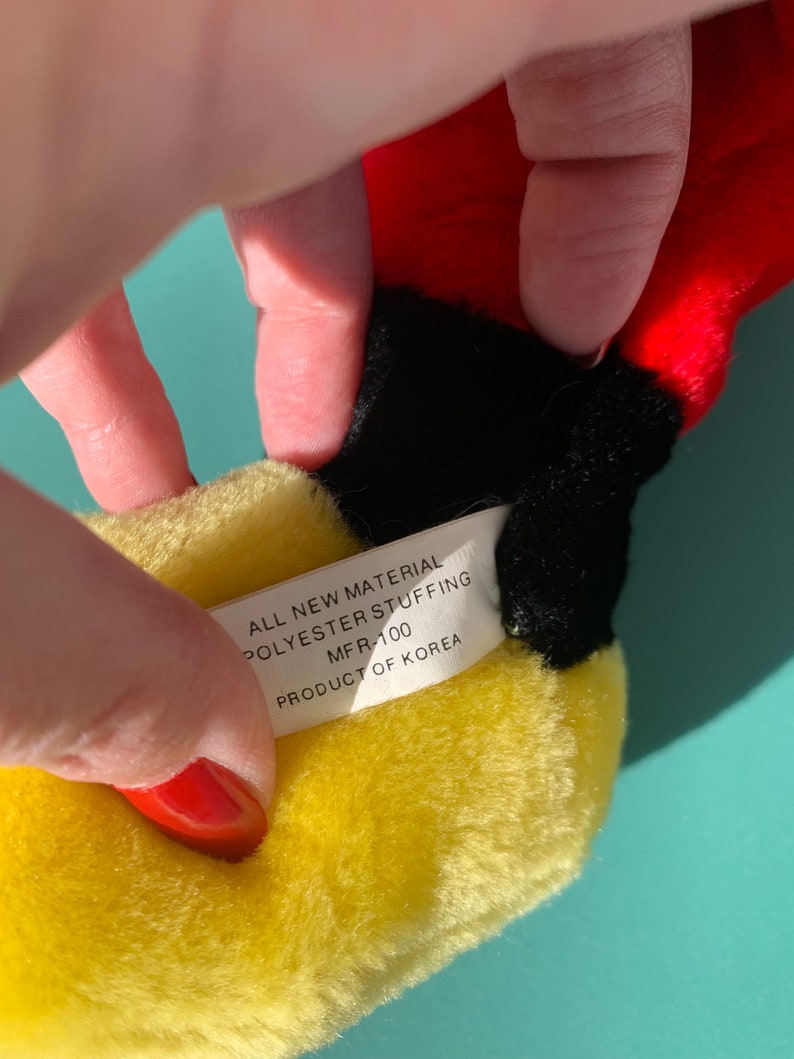 Puede incluir: Primer plano de un juguete de peluche amarillo con una etiqueta blanca que dice "All New Material Polyester Stuffing MFR-100 Product of Korea".