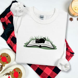 本の上に横たわるかわいい猫と花の刺繍入りスウェットシャツ、花と猫の刺繍入りパーカー、本好きのTシャツ、クルーネックスウェットシャツ。