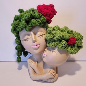 May include: A pair of ceramic planter heads with green crocheted hats and red flower accents. The heads are hugging each other.