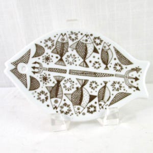 May include: White, fish-shaped decorative plate with a brown fish and trident design. The plate is supported by a clear stand. The fish design includes various fish, stars, and trident elements. The plate is likely ceramic.