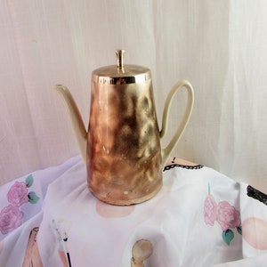 Op de afbeelding: Een vintage koffiepot met een metallic gouden afwerking. De pot heeft een hoge, cilindrische body met een gebogen handvat en een tuit. Het deksel is ook goudkleurig en het handvat en de tuit zijn gebroken wit. De pot staat op een witte stof met bloemenpatroon.