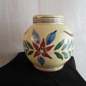 May include: A cream-colored ceramic vase with a rounded body and a short neck. The vase features hand-painted floral motifs in red, blue, and green, with horizontal blue and red stripes encircling the rim and the base of the vessel.