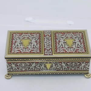 May include: A rectangular decorative box with ornate detailing. The box features a silver and gold color scheme with red accents. The top and sides are adorned with cherub and scrollwork designs. The box rests on four small, gold-colored feet.