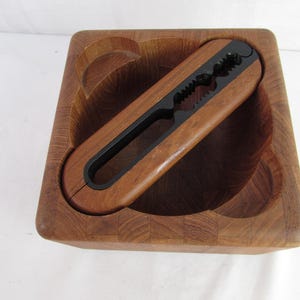 May include: A wooden nutcracker set featuring a square bowl with curved cutouts and a matching nutcracker. The nutcracker is brown with a black metal cracking mechanism. The set is made of wood.