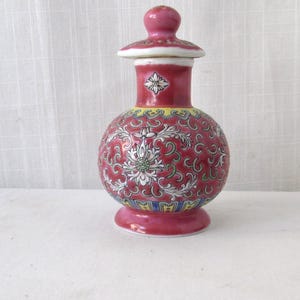 May include: A decorative, red ceramic vase with a lid. The vase features a bulbous body and a slender neck, adorned with intricate floral patterns in white, green, and yellow. The base and lid are a matching shade of red.