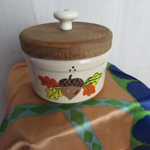 Puede incluir: Un tarro de cerámica color crema con tapa de madera y pomo blanco. El tarro está decorado con una bellota pintada y hojas coloridas. El tarro está sobre una superficie satinada.