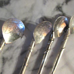 May include: Four silver-toned metal spoons with long, slender handles wrapped in a spiral wire design. The bowls of the spoons feature engraved floral and leaf patterns on their surfaces, with a polished metallic finish reflecting light.