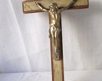 May include: A wooden crucifix with a gold-colored Jesus figure. The cross has a light-colored inlay with a brown border. The top of the cross has a small plaque with an inscription. The background is a white surface.