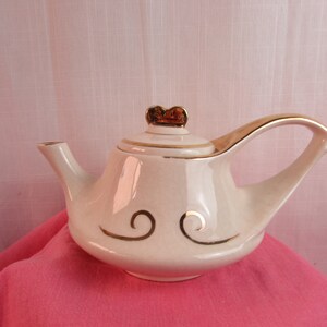 May include: A white teapot with a gold lid and gold swirl designs. The teapot has a unique, curved spout and handle.