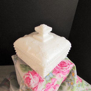 Puede incluir: Un plato de dulces de vidrio de leche blanco con una base cuadrada y una tapa abovedada. El plato está decorado con un diseño floral en relieve y tiene un borde de cuentas.