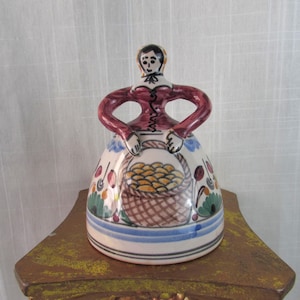 May include: A ceramic figurine of a woman in a dress, holding a basket. The figurine has a red bodice and arms, a white skirt with floral and basket designs, and a blue stripe at the base. The basket is filled with yellow objects.