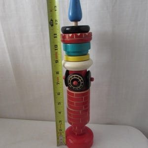 Könnte beinhalten: Ein vertikales Holz-Stapelspielzeug mit rotem Sockel und ziegelgemusterter Säule, gekrönt von gelben, türkisfarbenen, roten und schwarzen Ringen sowie einem blauen Kegel. Ein gelbes Maßband zeigt eine Höhe von etwa 39,4 cm an.