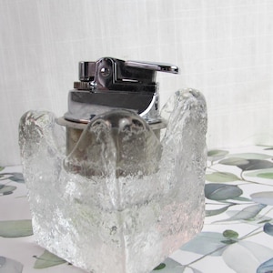 May include: A chrome lighter sits in a clear, textured glass holder. The glass holder has a unique, abstract design. The lighter is silver and has a simple, functional design. The background is a floral pattern.