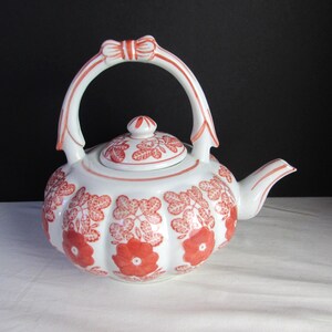 Può includere: Teiera in ceramica bianca con motivo floreale rosso. La teiera ha un corpo arrotondato, un manico curvo con dettaglio a fiocco e un coperchio abbinato. Il design floreale comprende grandi fiori e motivi a foglie. Il beccuccio è curvo.