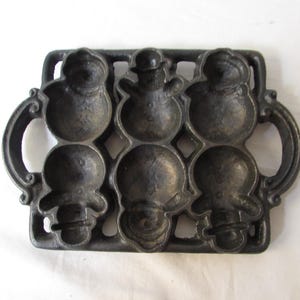 May include: A cast iron snowman-shaped pan with six moulds. The pan is dark grey with a rectangular shape and handles on each side. Each mould is a snowman with a hat, arms, and a round body. The pan is designed for baking.