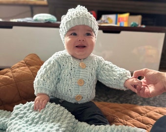 Puede incluir: Un bebé con gorro y cárdigan de punto azul claro con botones de madera. El bebé está sentado sobre una manta de punto azul claro. El cárdigan y el gorro son hechos a mano.