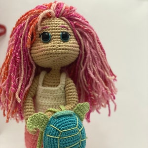 May include: A crocheted doll with pink and red yarn hair, wearing a white crocheted dress and holding a blue and green crocheted turtle.