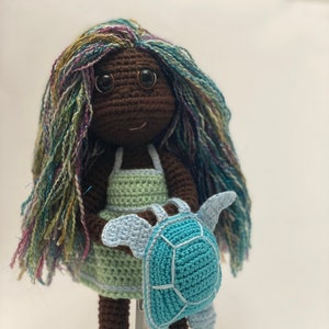 May include: A crocheted doll with brown skin, green and blue hair, and a green dress. The doll is holding a blue crocheted turtle.