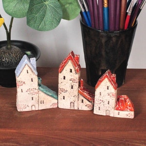 Puede incluir: Una colección de cuatro casas en miniatura de cerámica con detalles en rojo, blanco y azul. Las casas están dispuestas en fila sobre una superficie de madera. Hay una pequeña planta y una figura de un personaje de dibujos animados en el fondo.