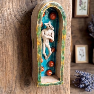 May include: A ceramic wall hanging featuring an arched frame with a textured beige exterior and a vibrant blue interior. Inside the arch, a sculpted white figure with a fox-like head stands with arms crossed, surrounded by small, round red spheres.