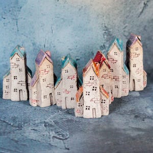 Puede incluir: Un conjunto de pequeñas casas de cerámica hechas a mano con tejados coloridos. Las casas están pintadas de blanco con detalles de ventanas y puertas. Los tejados son azules, morados y rojos. Dispuestas sobre una superficie azul texturizada.