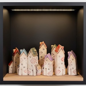 Könnte beinhalten: Eine Sammlung kleiner, handbemalter Keramikhäuser mit roten, blauen, grünen und violetten Dächern. Die Häuser stehen auf einem Holzregal in einem schwarzen, eingelassenen Display. Die Häuser haben ziegelartige Details.