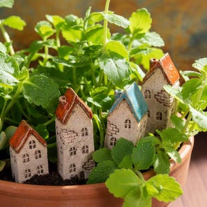 Puede incluir: Una maceta de terracota llena de plantas verdes y casas de cerámica en miniatura. Las casas tienen paredes blancas, detalles de ladrillo y tejados naranjas o azules. Una planta suculenta es visible en el lado izquierdo.