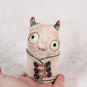 May include: A handmade ceramic sculpture of a whimsical creature with large green eyes, small horns, and a stitched garment. The sculpture is primarily off-white with brown and green accents, and black button details.
