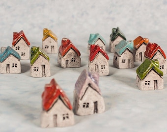 Peut inclure: Une collection de petites maisons en céramique avec des toits colorés. Chaque maison a un extérieur blanc avec une porte et deux fenêtres. Les toits sont peints de différentes couleurs, dont le rouge, le vert, le bleu et le jaune. Les maisons sont disposées sur une surface bleu clair.