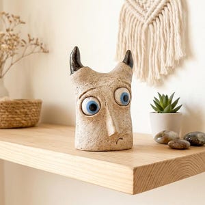 Può includere: Una scultura in ceramica beige raffigurante una creatura dalla superficie ruvida, con due corna marrone scuro e due grandi occhi rotondi blu brillante. La figura presenta un naso pronunciato e una piccola linea curva che forma la bocca.