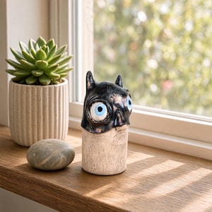 May include: A small ceramic sculpture featuring a black, glossy, horned creature head with large, wide, light blue eyes, resting on a textured, cylindrical, off-white base. The object has a smooth, reflective finish on the upper portion.