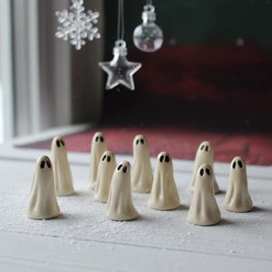 Può includere: Una collezione di piccole statuette di fantasmi in ceramica bianco sporco con ritagli di occhi neri. I fantasmi sono disposti su una superficie bianca, con ornamenti decorativi appesi sullo sfondo. Il tema è Halloween o decorazioni spettrali.
