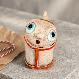 May include: A small, cylindrical ceramic figurine with a textured, off-white surface. It features two large, prominent eyes with bright blue irises and black pupils, a small nose, and a tiny, open red mouth. An orange-red glazed trim outlines the face.