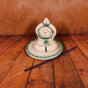 Puede incluir: Un quemador de incienso de cerámica con forma de figura en posición de meditación sentada con las manos levantadas sobre la cabeza. La figura es de color blanco roto con detalles en turquesa moteado en la base y la cabeza. Un incienso morado.