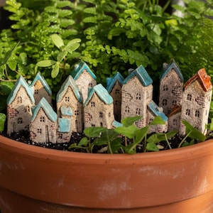 Puede incluir: Una maceta de terracota llena de vegetación, que alberga un pueblo en miniatura de casas de cerámica. Las casas tienen paredes de color beige, tejados azules o naranjas y pequeñas ventanas, creando una escena de jardín caprichosa.