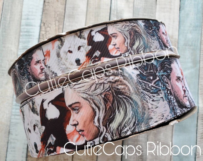 May include: Two rolls of white and brown ribbon with a printed design of characters from the TV show Game of Thrones. The ribbon features images of Daenerys Targaryen, Jon Snow, and a direwolf.