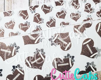 May include: White ribbon with a brown glitter football heart pattern. The ribbon has silver glitter bows and the text "Cutie Caps Ribbon" in pink.