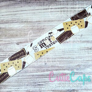 May include: White ribbon with a brown and gold print of s'mores. The ribbon has the text "Official S'more Tester" printed on it.