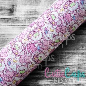 May include: A roll of pink and white faux leather fabric with a repeating pattern of cartoon cats in spaceships, planets, stars, and crystals. The fabric has a textured surface and the words "CutieCaps Ribbon" are visible in the lower right corner.