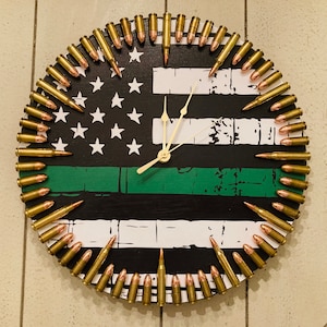 May include: A round clock with a black background and a green stripe. The clock face features a US flag design with white stars on a blue field and red and white stripes. The clock hands are white and the numbers are replaced with bullet casings.