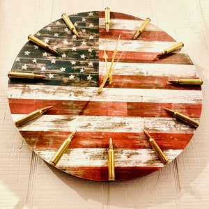 May include: A round clock with a distressed American flag design. The clock hands are gold, and the numbers are represented by gold bullet casings.