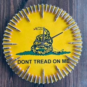 May include: A yellow clock face with a coiled snake and the words "Don't Tread On Me". The clock face is surrounded by a ring of bullet casings.