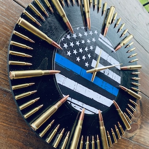 May include: A black clock face with a thin blue line and American flag design. The clock hands are gold. The numbers are made of bullet casings.