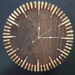 May include: A round wooden clock with a dark brown wood face and gold-colored hands. The clock is decorated with a circle of bullet casings around the edge.