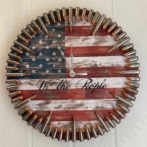 May include: A round clock with a distressed American flag design. The clock face is surrounded by bullet casings. The text "We the People" is printed on the clock face.