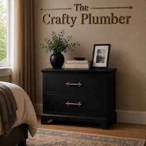 May include: A black two-drawer bedside table with copper-coloured handles. The table is adorned with a black vase, books, and a framed picture. The wall above features the text "The Crafty Plumber" in gold lettering.